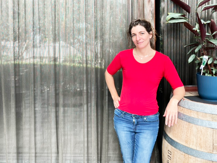 A woman in a red top and jeans stands beside a barrel and smiels at the camera
